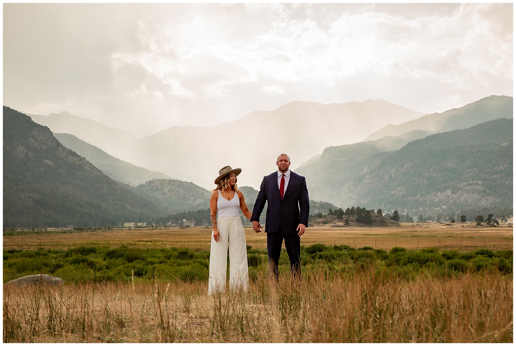 Bride and groom in a field on a partly cloudy day holding hands with the rocky mountains in the background